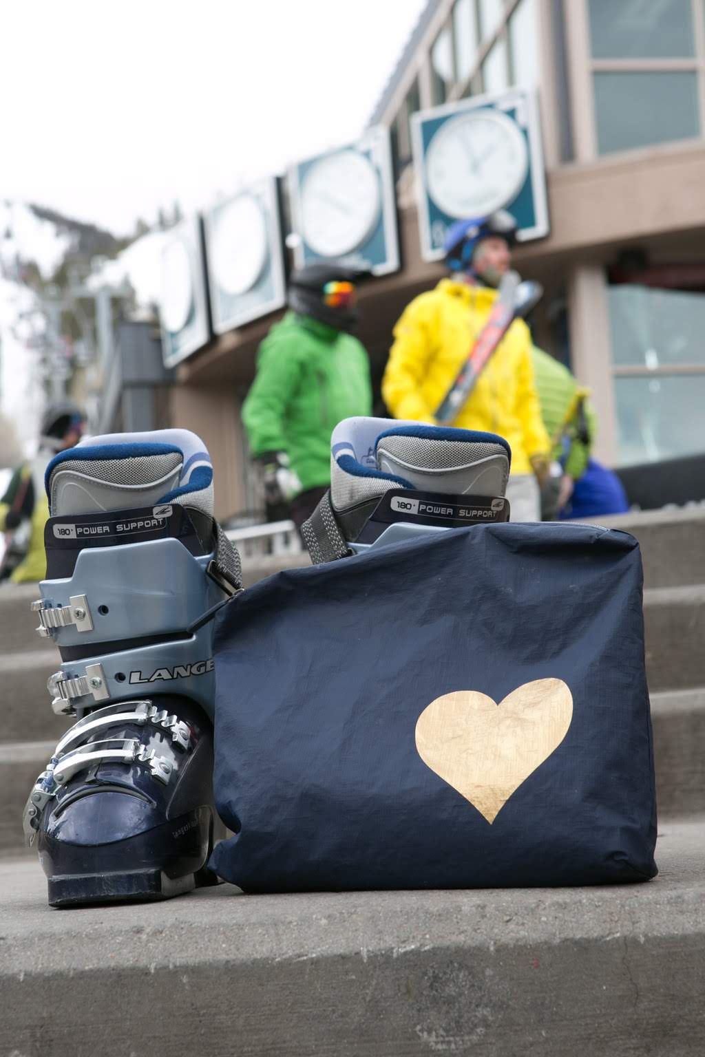 Navy bag with a gold heart design on steps, with people and ski equipment in the background.