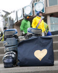 Navy bag with a gold heart design on steps, with people and ski equipment in the background.