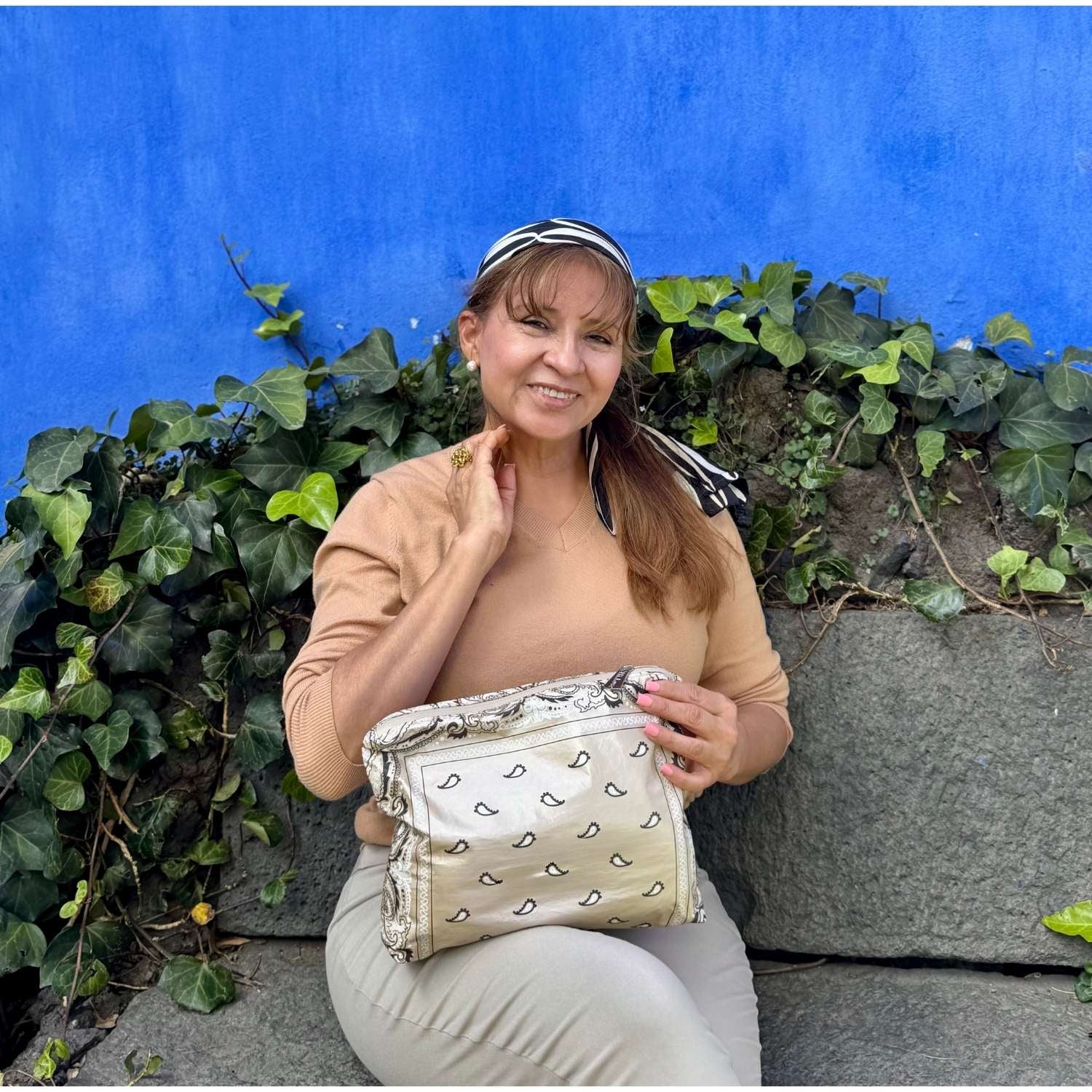 Women carrying beige bandana print clutch sitting on a bench with greenery and a bright blue wall