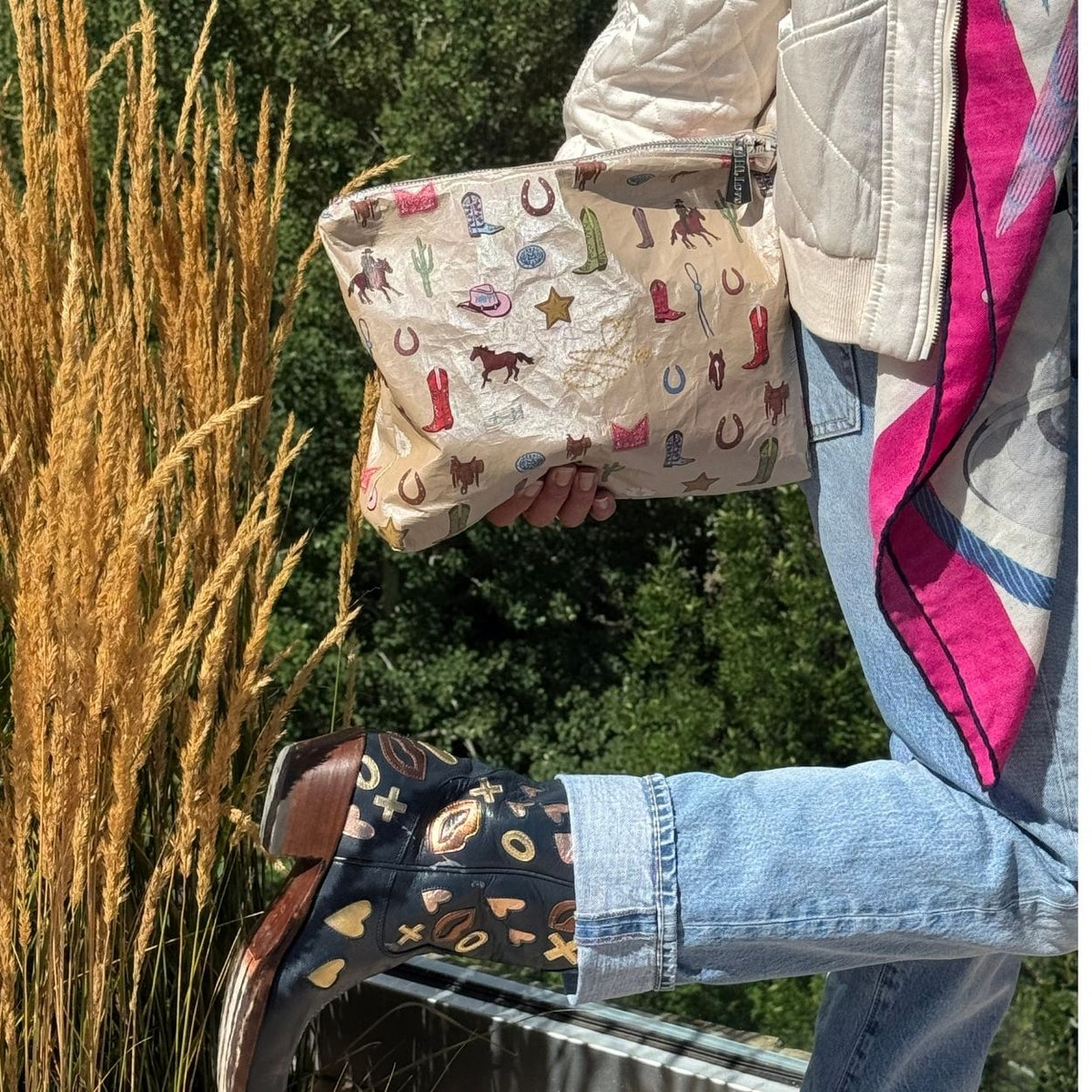 Hand holding a beige pouch with western boots, hats, horse and cactus on it in an outdoor setting with colorful cowboy boots worn on a person.