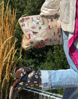 Hand holding a beige pouch with western boots, hats, horse and cactus on it in an outdoor setting with colorful cowboy boots worn on a person.