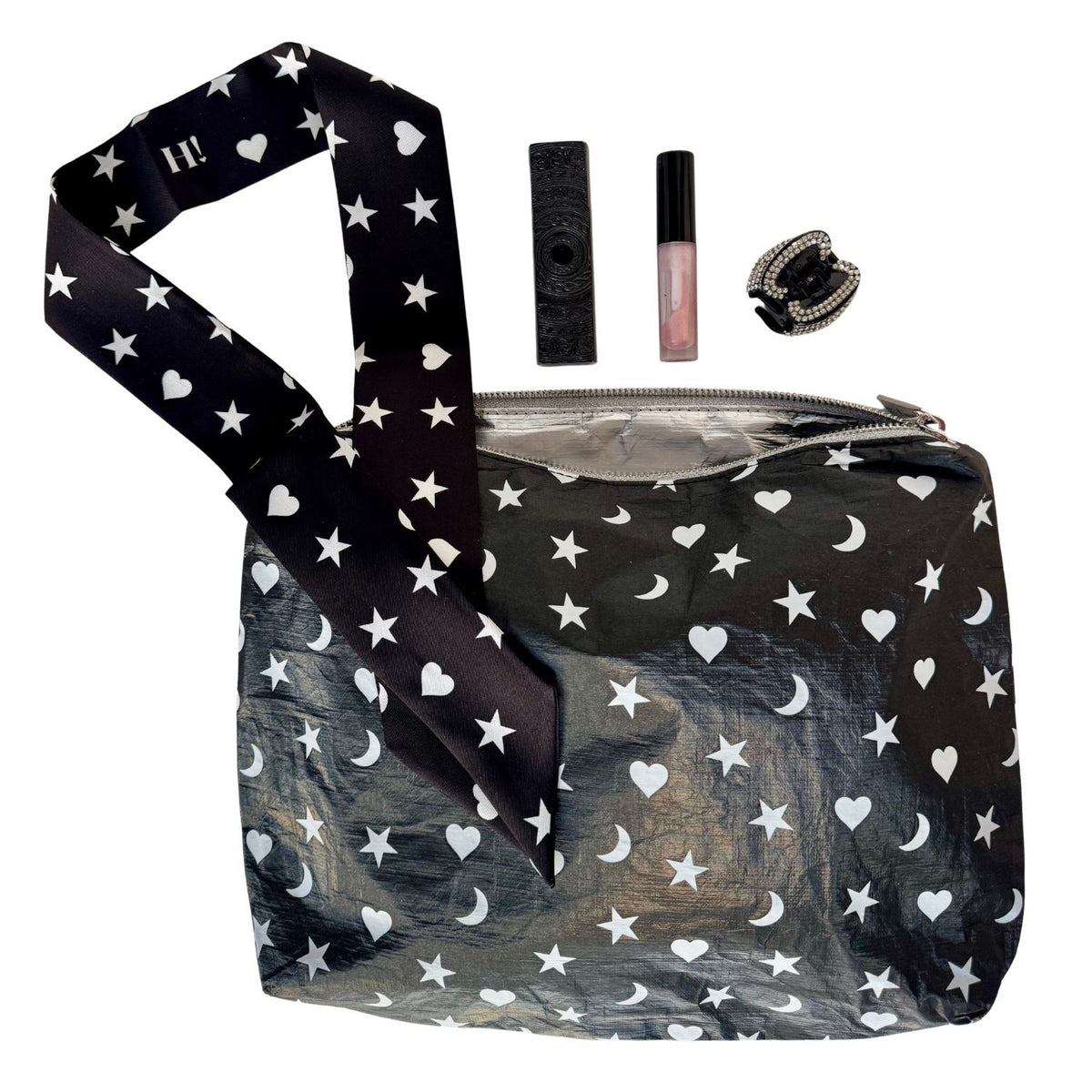 A black zippered cosmetic pouch with a white star, moon, and heart pattern, shown with a matching patterned silk scarf wristlet strap and small accessories perfume,  lip gloss, and a black rhinestone hair clip on a white background.
