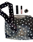 A black zippered cosmetic pouch with a white star, moon, and heart pattern, shown with a matching patterned silk scarf wristlet strap and small accessories perfume,  lip gloss, and a black rhinestone hair clip on a white background.
