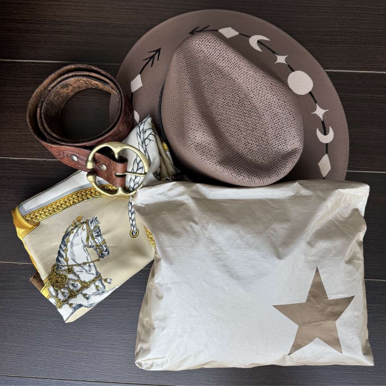 Cowboy hat, belt, silk scarf with horse design, and star-patterned bag on a wooden surface