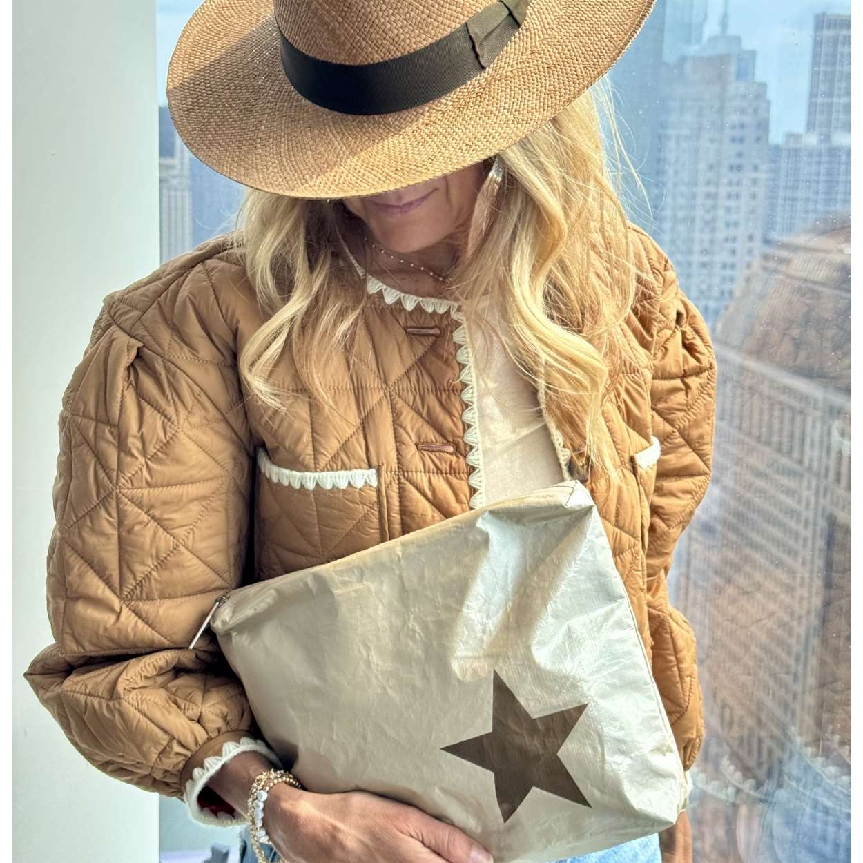 Person wearing a quilted jacket and straw hat, holding a beige bag with a star design.