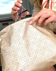 A close-up of a person holding a large beige tote bag with a repeating geometric monogram pattern. The person is wearing a beige coat and holding sunglasses in one hand while reaching toward the bag with the other.