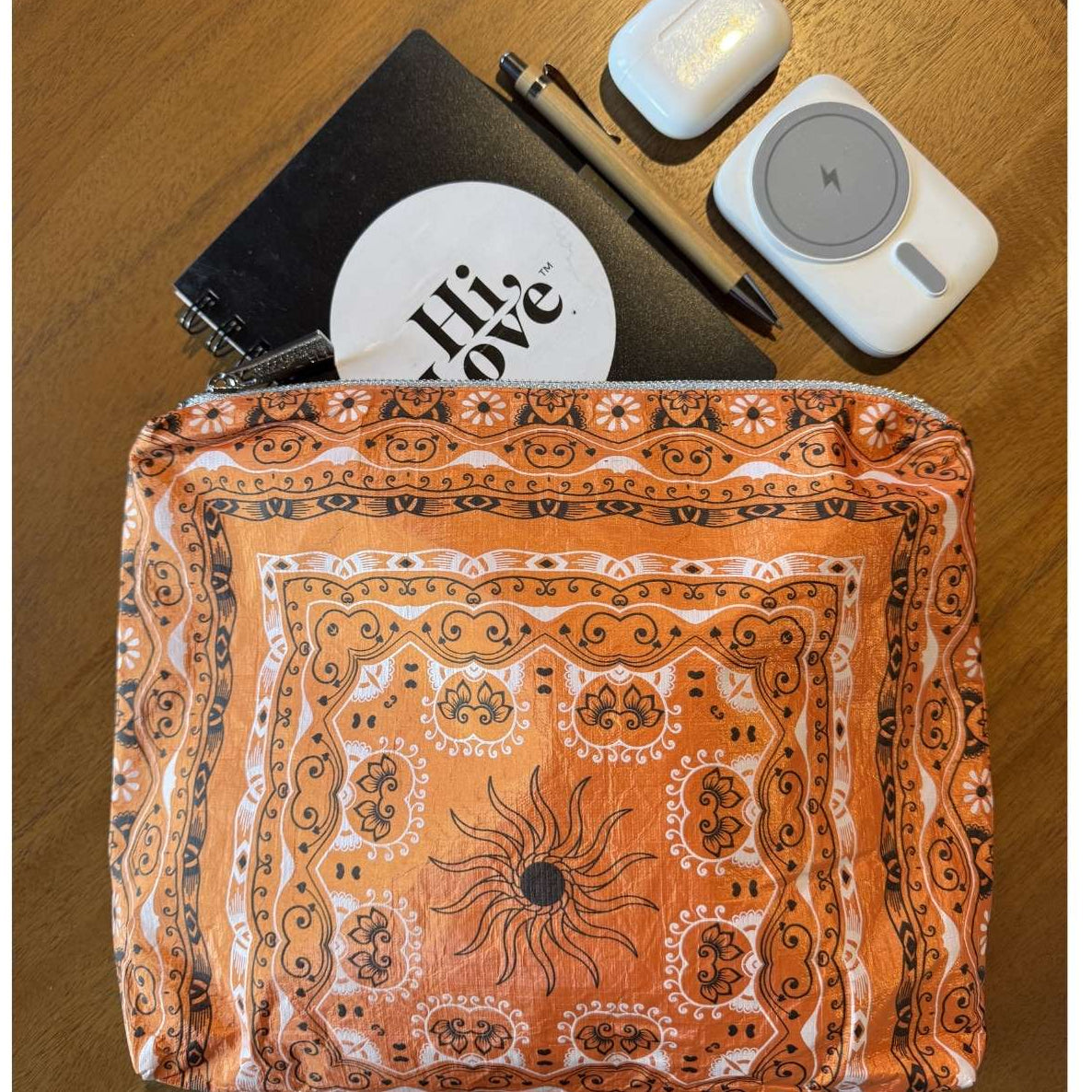 Zipper Pouch in Orange Bandana Print with notebook, pen, charger and ear buds on a wooden surface.
