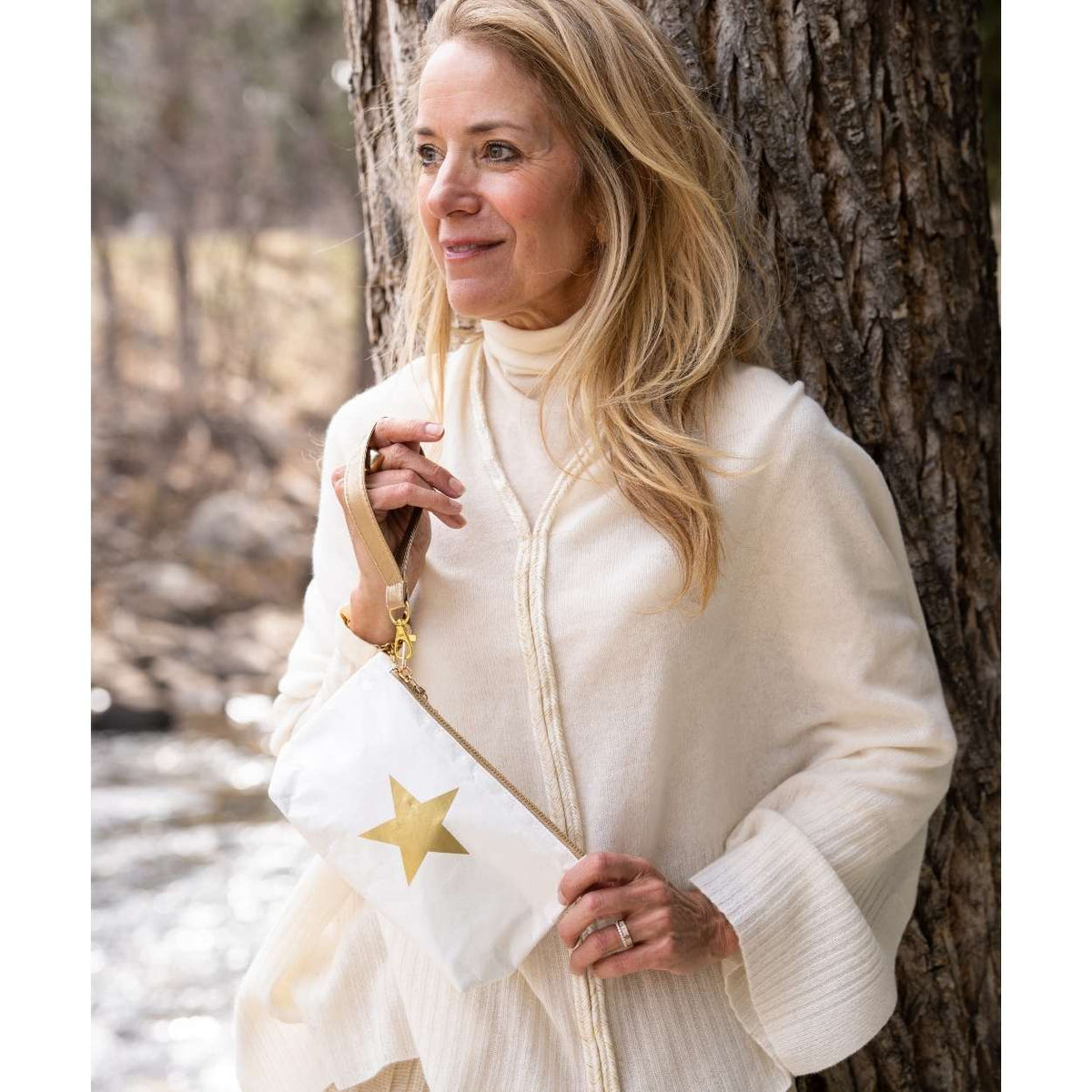 A woman standing outdoors next to a tree, wearing a cream ribbed turtleneck sweater and holding a white zippered pouch bag with a gold star detail.