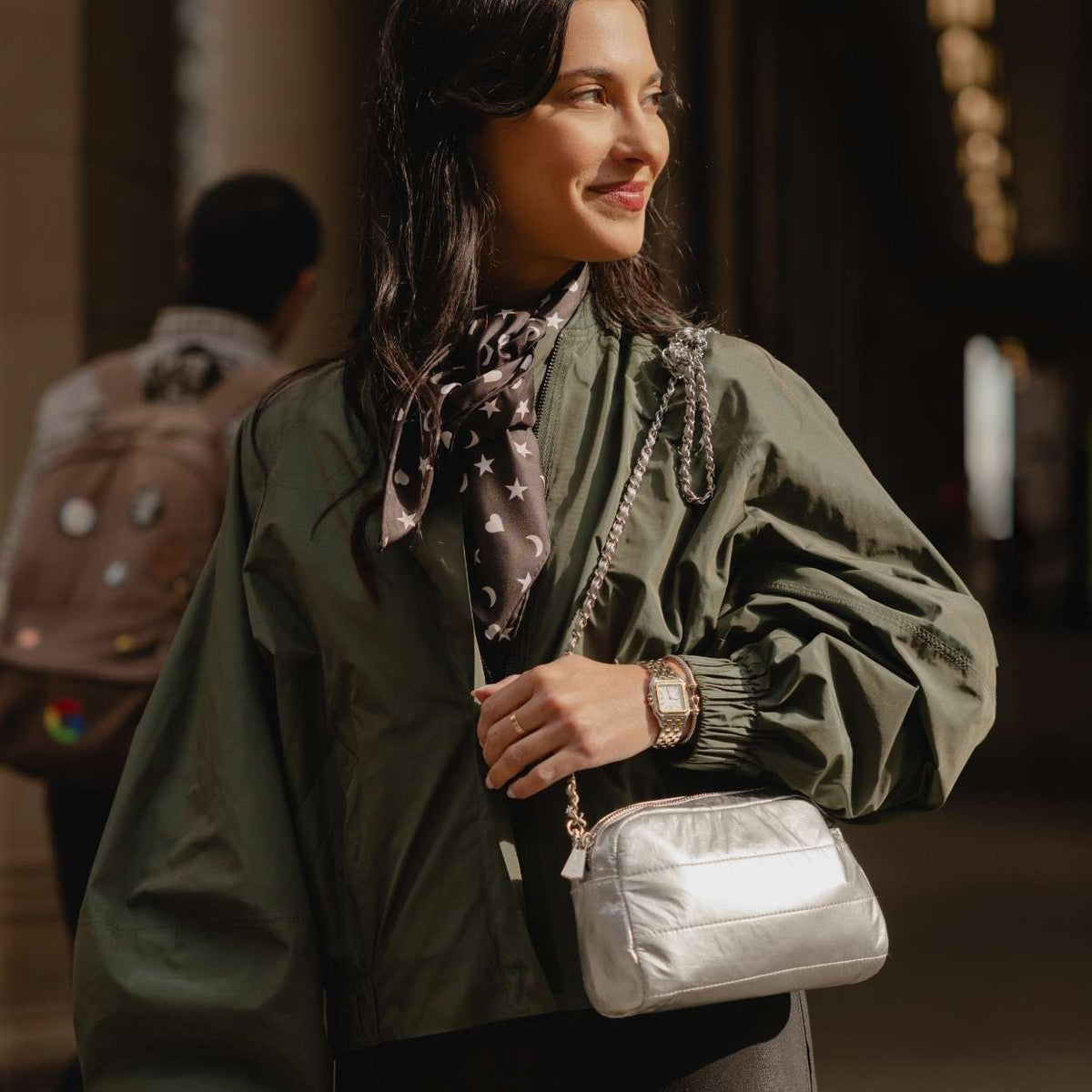 A woman wearing an olive green bomber jacket and a patterned scarf, carrying a shiny silver crossbody bag with a chain strap in a dimly lit indoor setting.