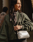 A woman wearing an olive green bomber jacket and a patterned scarf, carrying a shiny silver crossbody bag with a chain strap in a dimly lit indoor setting.