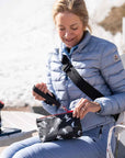 A woman sitting outdoors by a beach or lake wearing a light blue puffer jacket and blue jeans, opening a black zip pouch with white skier silhouettes. Another matching black and white pouch rests on a bench beside her.