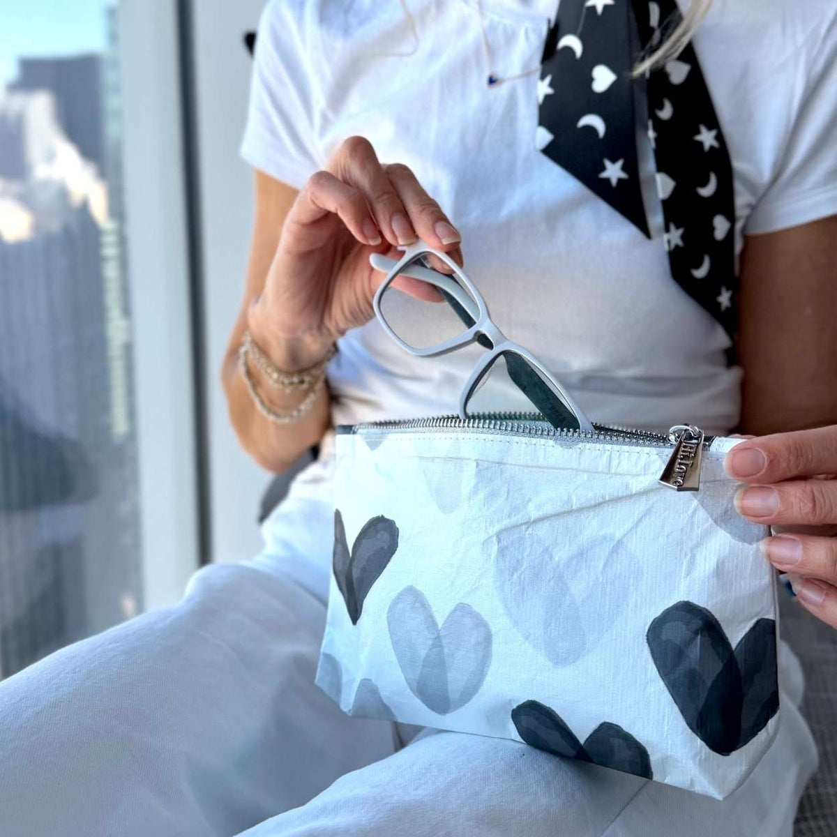 A person holding a white zippered pouch with a black and gray heart pattern, holding a pair of eyeglasses inside. The person is wearing a white t-shirt and white pants.