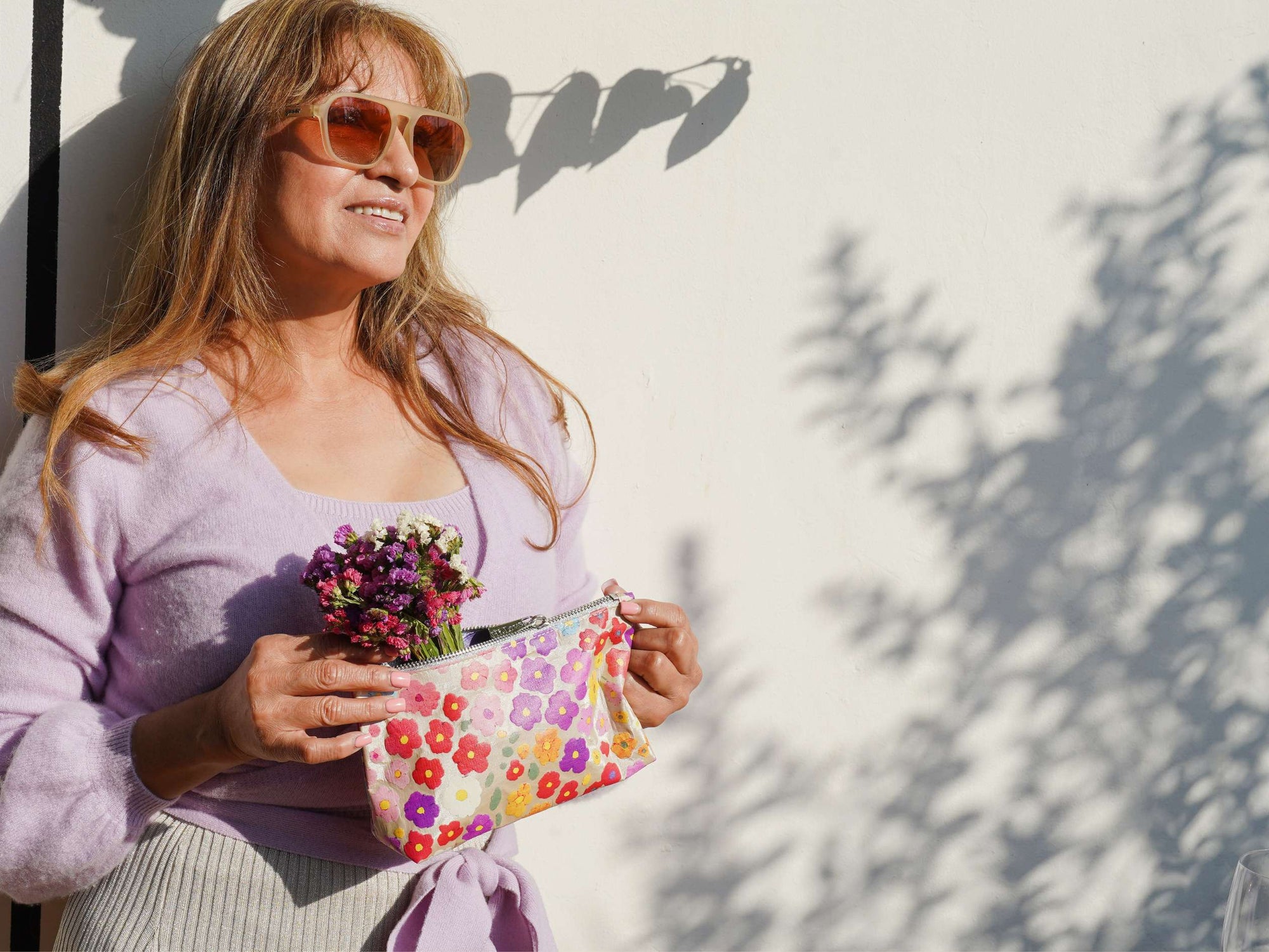 Woman holding a colorful floral clutch and flowers against a white wall with shadows.