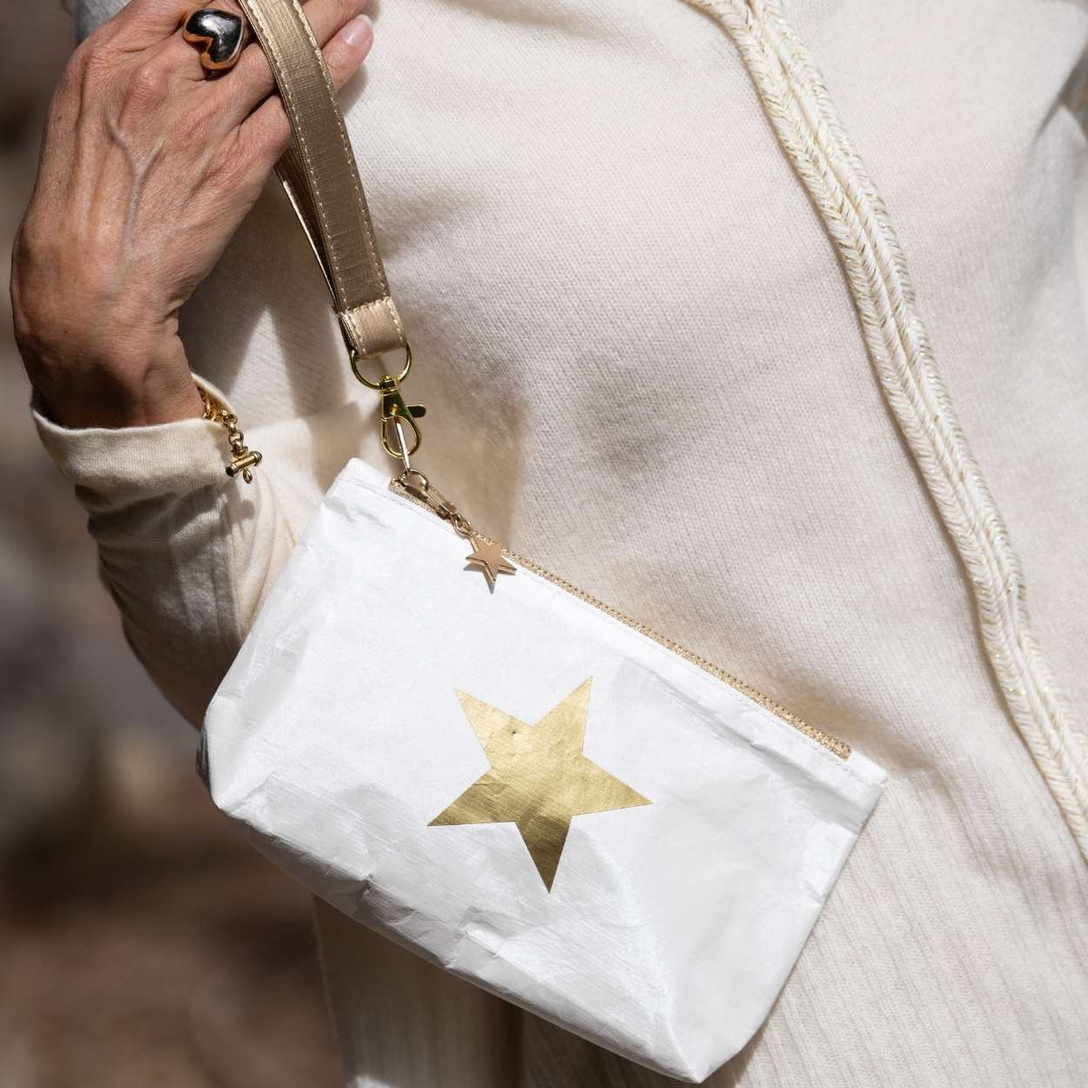A close-up of a person wearing a cream knit sweater and holding a small white crossbody pouch with a metallic gold star on the front and a gold-toned strap.