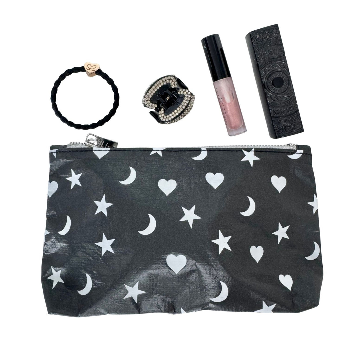 A black cosmetic pouch with a white celestial pattern of stars, hearts, and crescent moons, displayed alongside beauty accessories including a black braided hair tie, a rhinestone ring, a lip gloss tube, and perfume on a white background.