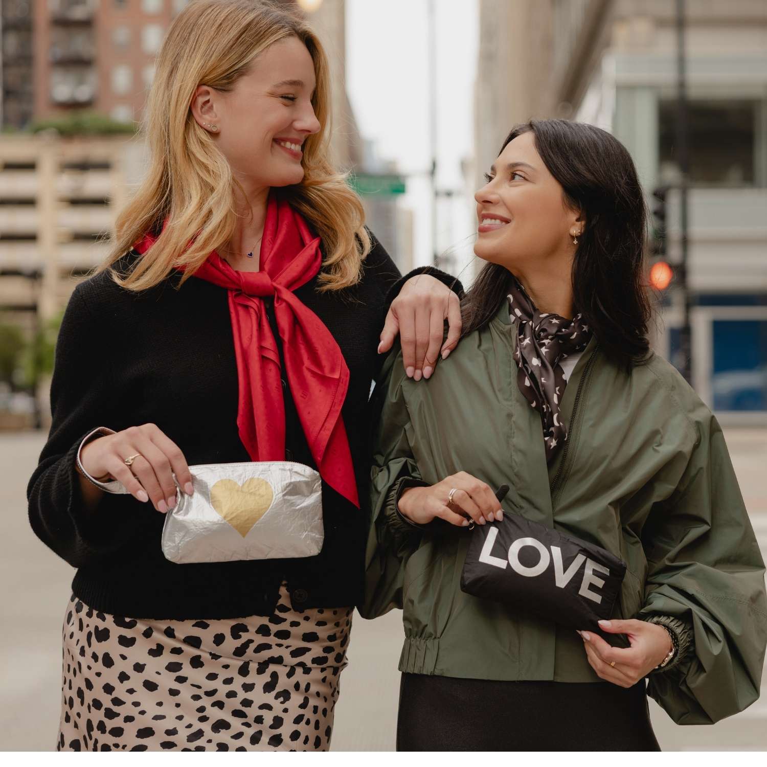 Two women standing outdoors on a city street, each holding a black clutch bag with Gold Heart and LOVE text. The woman on the left wears a black top and a bright red scarf, and the woman on the right wears an olive green jacket and a dark patterned scarf.