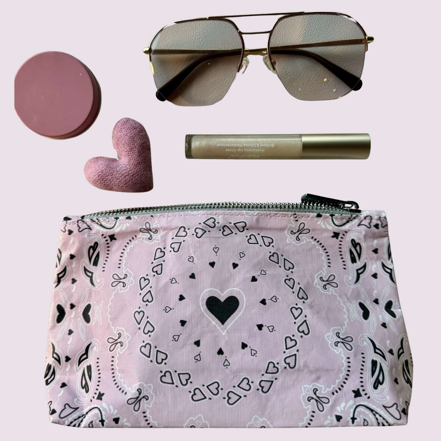 Makeup bag with bandana print heart pattern, sunglasses, pink heart-shaped compact, and  lip balm on a light pink background