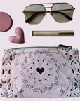 Makeup bag with bandana print heart pattern, sunglasses, pink heart-shaped compact, and  lip balm on a light pink background