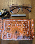 Orange bandana pouch with sun design, a bracelet, eye glasses and lip balm on a wooden surface