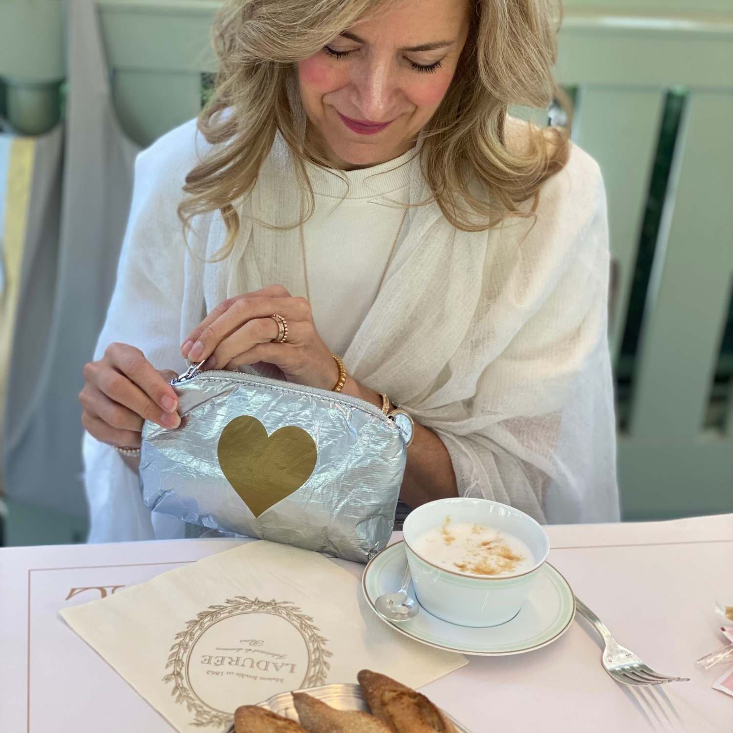 Woman in a white outfit with a gold heart pouch, sitting at a table with a cup of coffee and pastries.