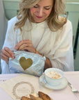 Woman in a white outfit with a gold heart pouch, sitting at a table with a cup of coffee and pastries.