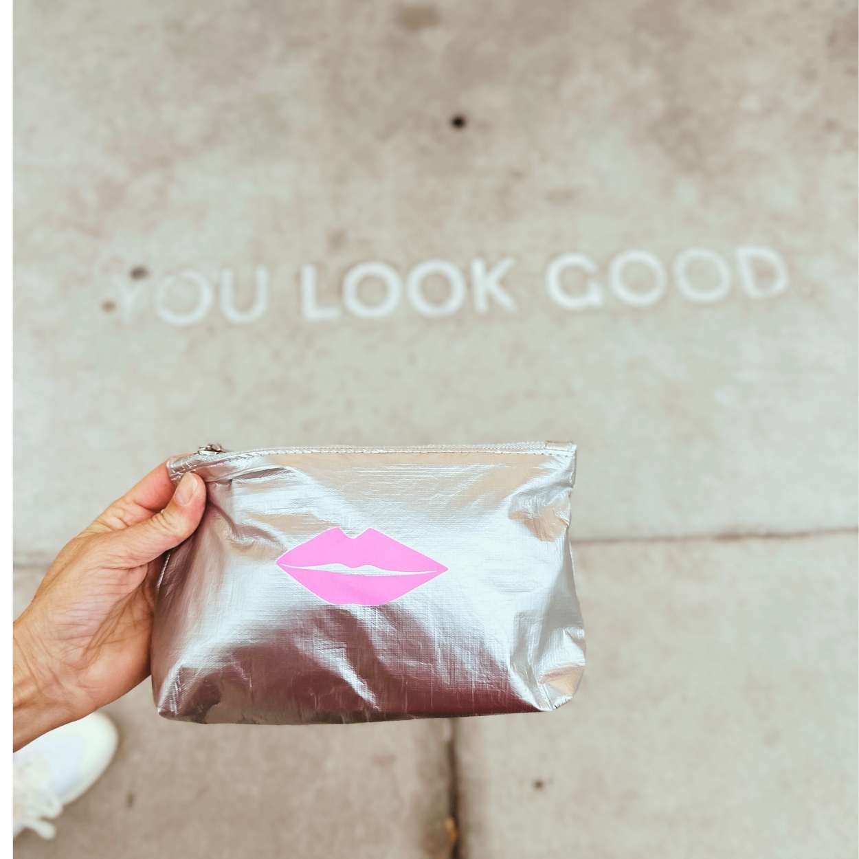 Hand holding a silver pouch with pink lips design against a neutral background with text that says "you look good".