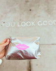 Hand holding a silver pouch with pink lips design against a neutral background with text that says "you look good".