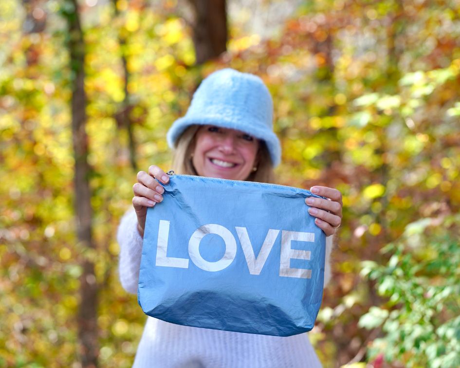 Woman smiling wearing blue hat holding blue pouch with "LOVE" on colorful leaf background