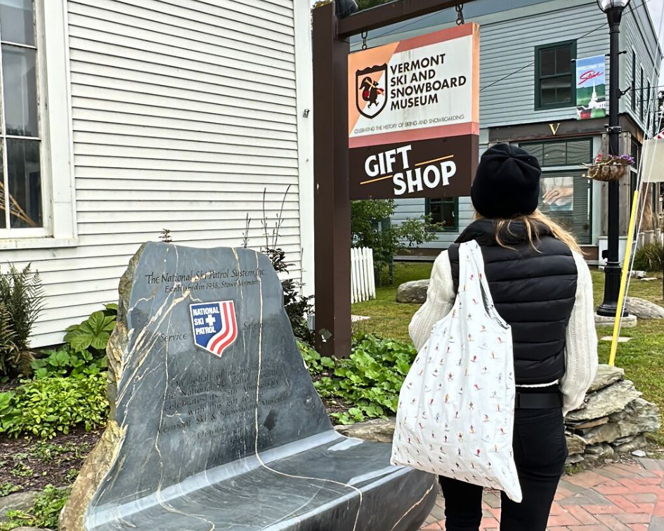 Person standing outdoors near Vermont ski and snow board museum gift shop holding carryall tote bag in dancing skiers pattern