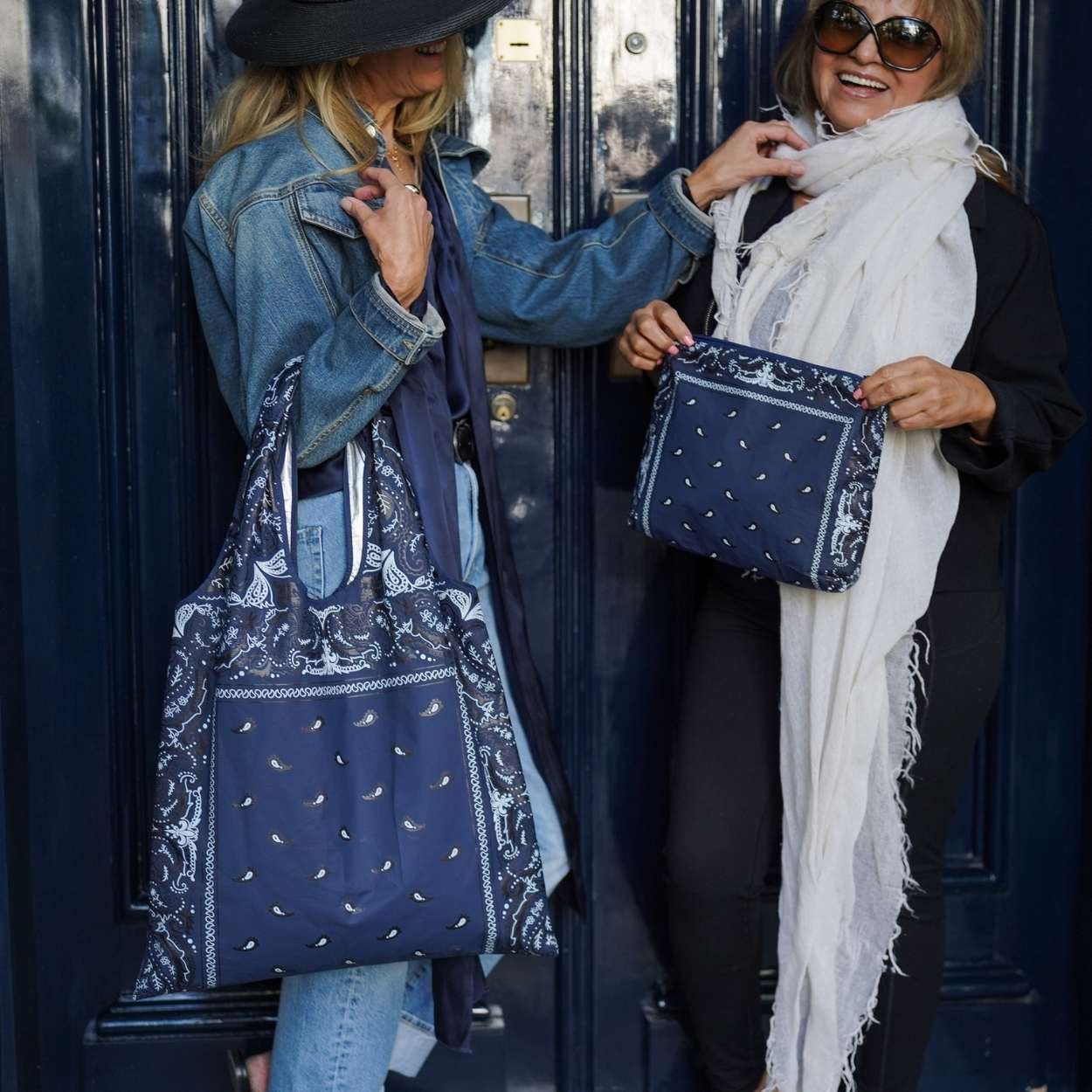 Two women holding blue bandana patterned tote bags against a navy blue door 