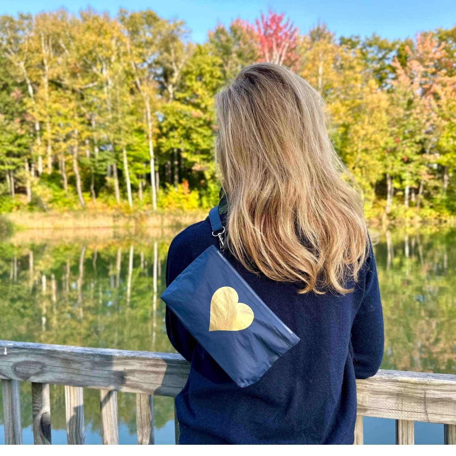 Woman wearing a navy blue wristlet with gold heart overlooking a lake and fall foliage
