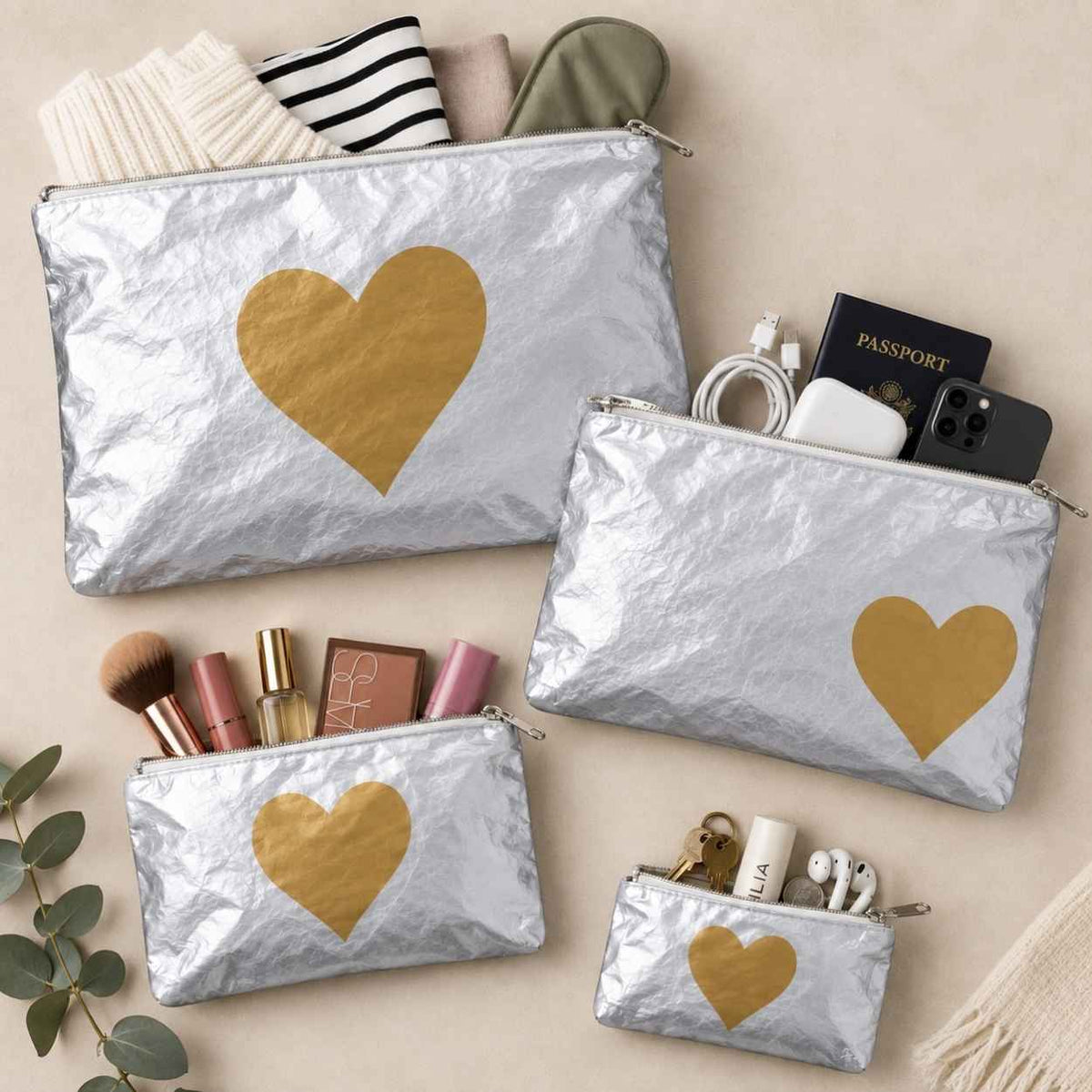 Set of four silver travel pouches with gold hearts on beige background holding various beauty and travel itrems