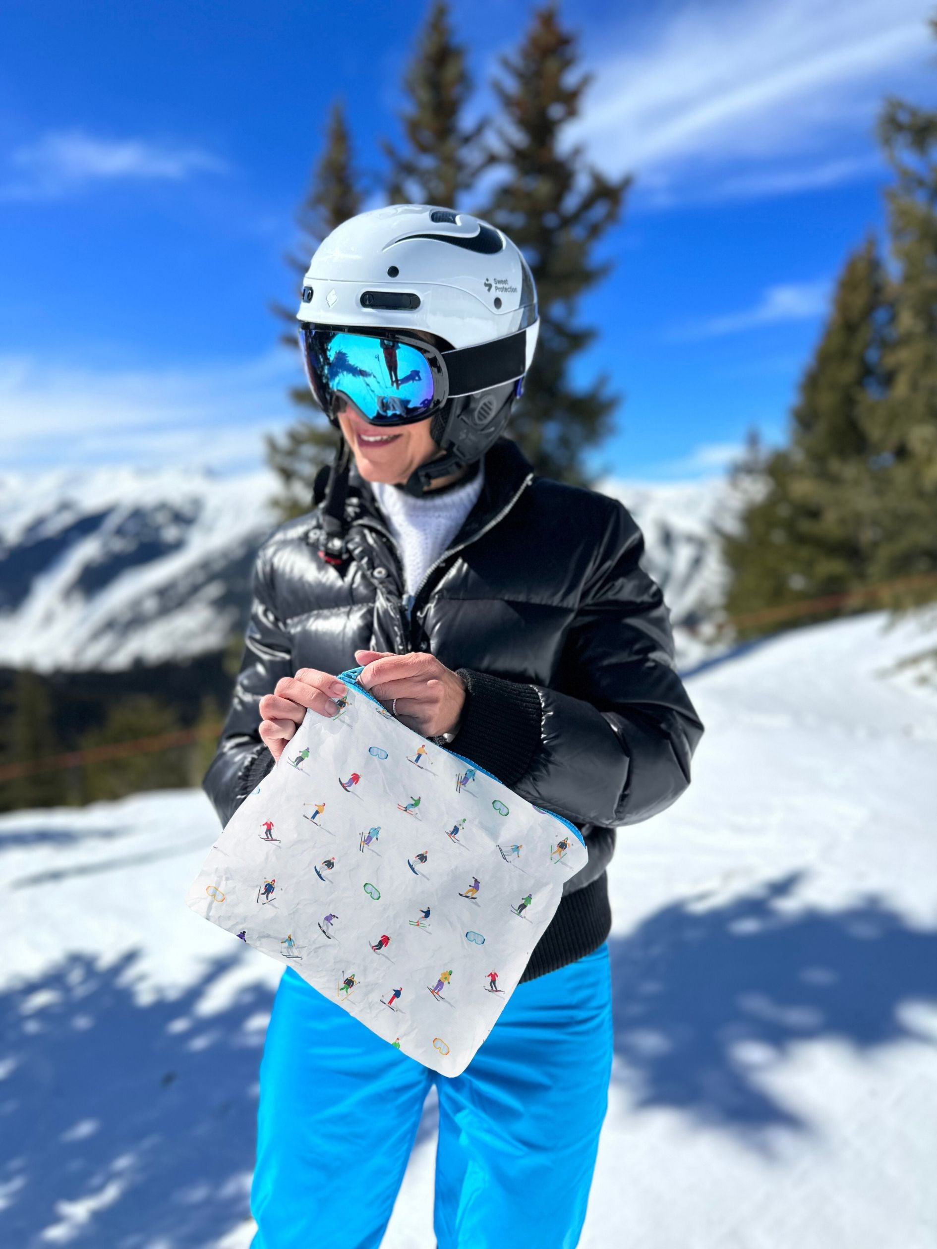 Skier on top of snow capped mountain holding a skier patterned zipper pouch