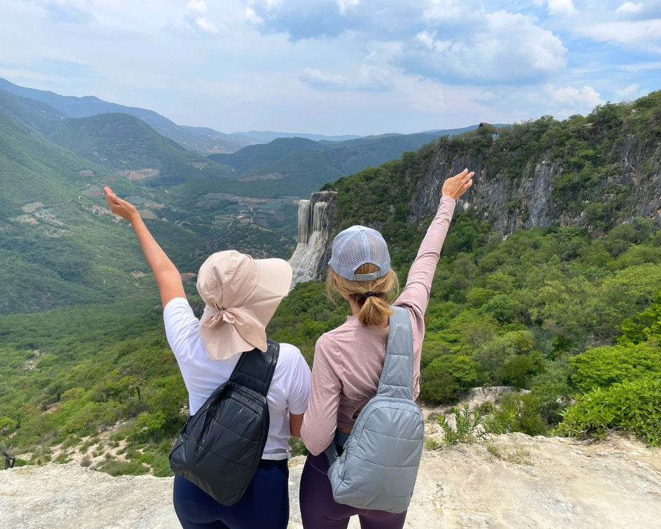 Two people wearing crossbody backpacks in cool gray and shimmer black, overlooking view with waterfall
