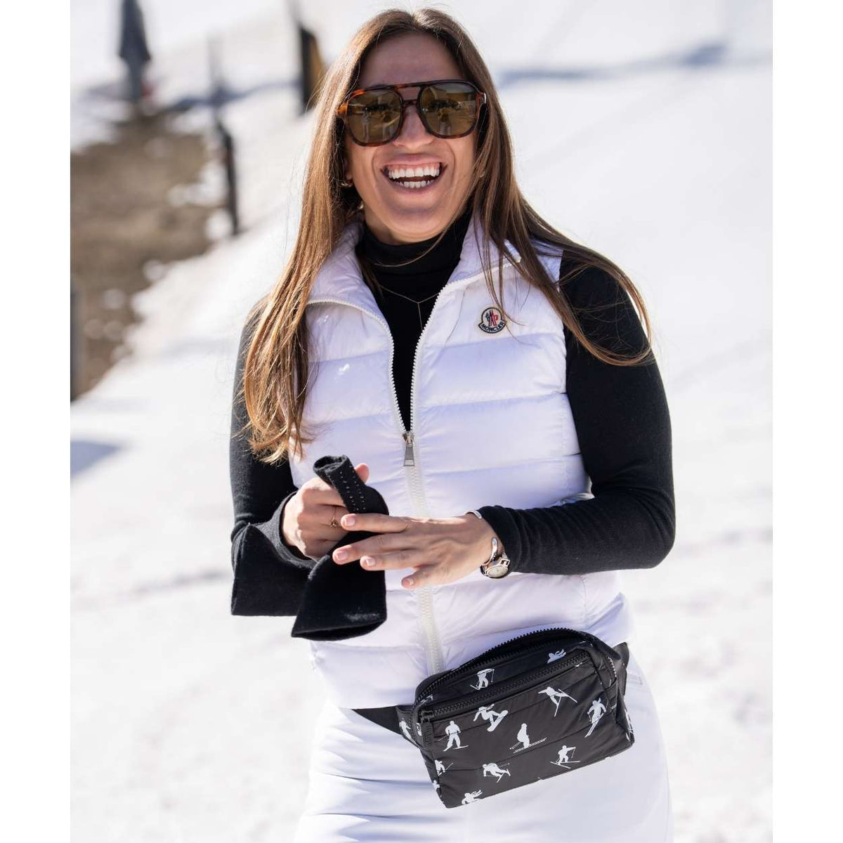A woman in a snowy outdoor setting wearing a white puffer vest over a black long-sleeve top, white ski pants, black sunglasses, and a black ski patterned fanny pack.