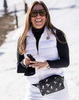 A woman in a snowy outdoor setting wearing a white puffer vest over a black long-sleeve top, white ski pants, black sunglasses, and a black ski patterned fanny pack.