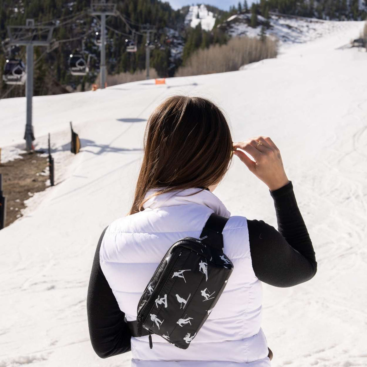 A woman on a ski slope seen from behind, wearing a black crossbody belt bag with a white ski pattern over a white puffer vest and black long-sleeve shirt.