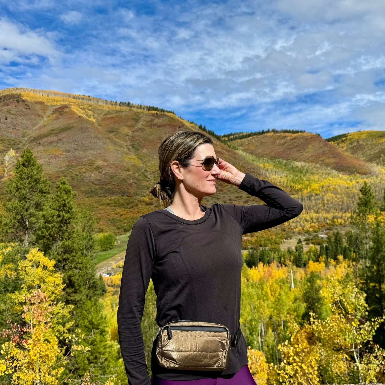Woman smiling wearing Puffer Crossbody Fanny Pack Belt Bag in Shimmer Bronze with fall mountain background