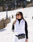 A woman wearing a white puffer vest over a black long-sleeve top, white ski pants, sunglasses, and a black ski patterned fanny pack stands on a snowy mountain slope with ski lifts in the background.