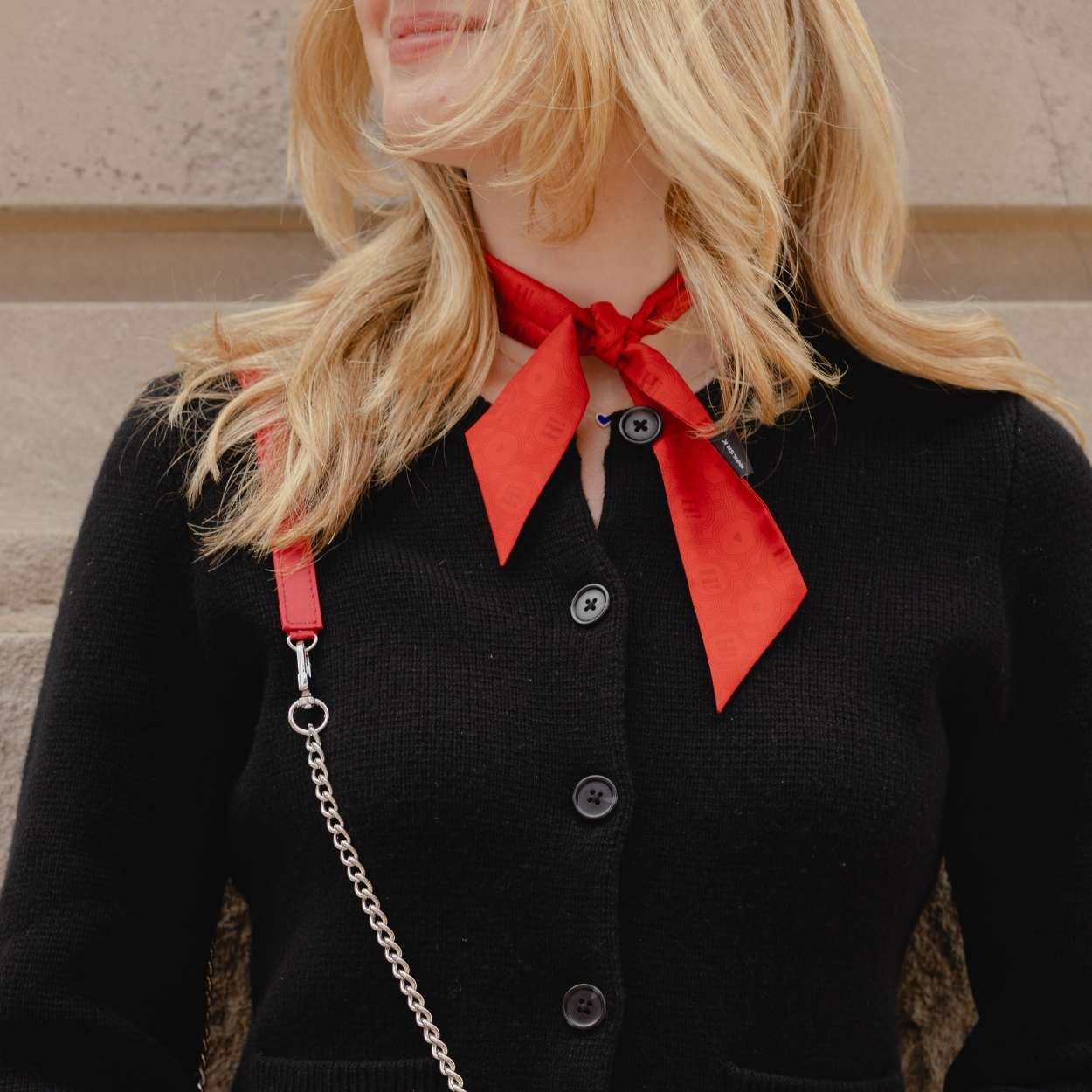 A close-up of a woman wearing a black button-front cardigan with a bright red satin bow tie at the collar, a red shoulder strap, and a silver chain strap, against a beige stone wall.