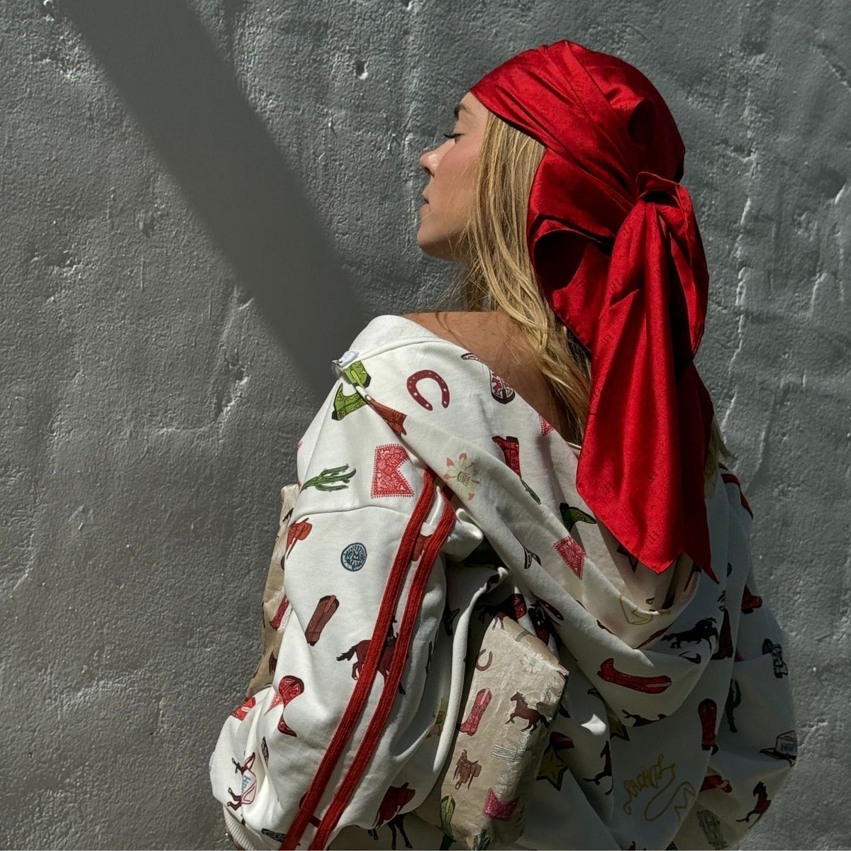 Woman wearing a western print zip up sweatshirt with a Red Silk head scarf standing against a white wall.