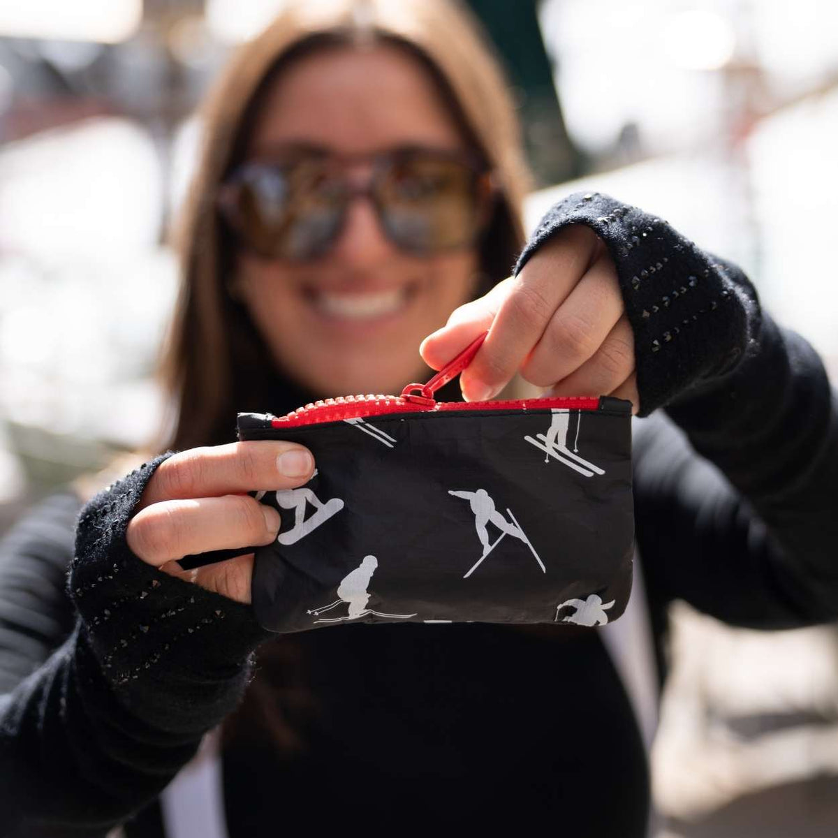 A person wearing sunglasses and a black knit sweater holds up a small black zip pouch with a red zipper and white ski-themed silhouette print.