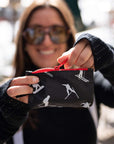 A person wearing sunglasses and a black knit sweater holds up a small black zip pouch with a red zipper and white ski-themed silhouette print.