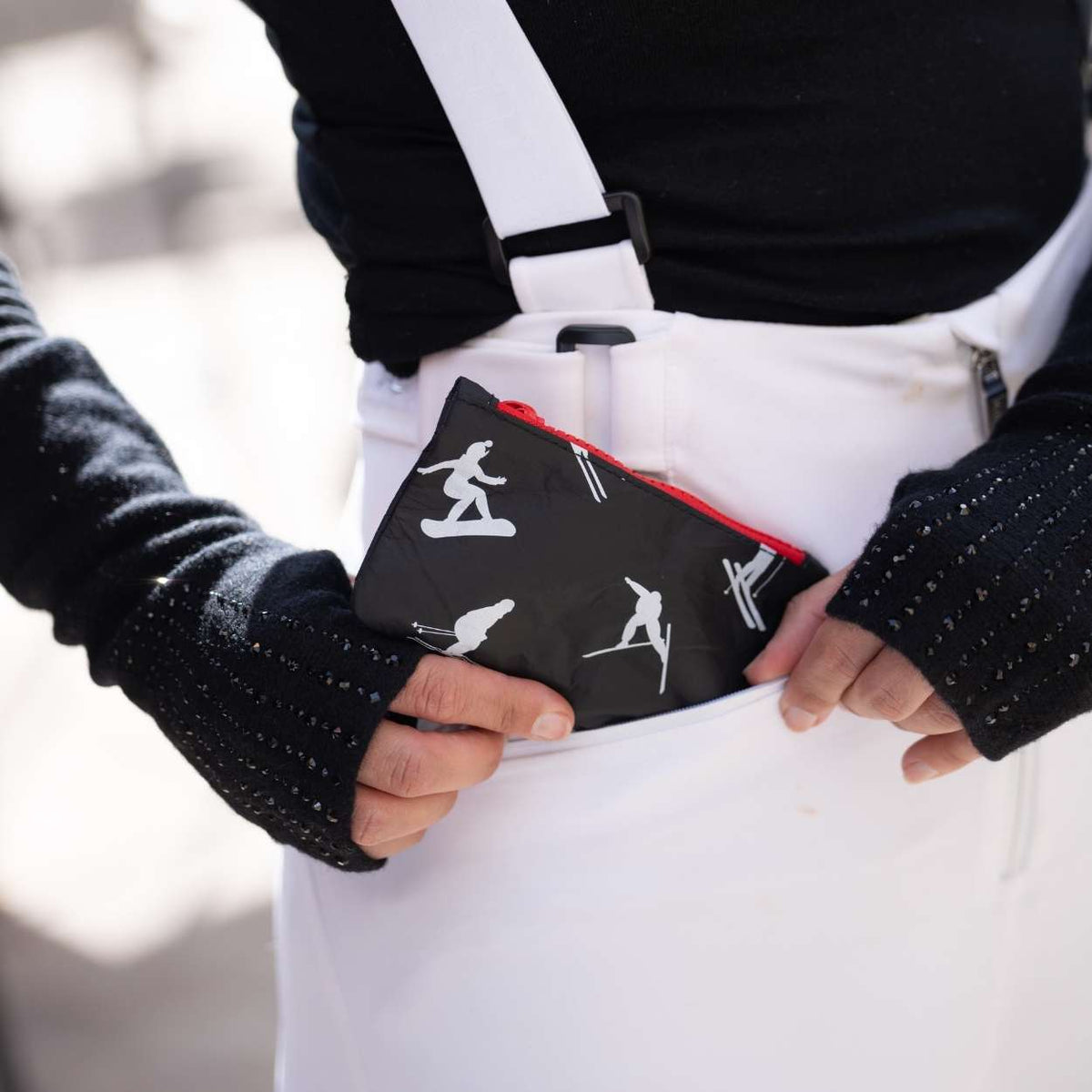 A close-up lifestyle shot of a person wearing white ski overalls and a black knit sweater, holding a black zippered pouch with white snowboarder and skier graphics in their pocket.