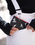A close-up lifestyle shot of a person wearing white ski overalls and a black knit sweater, holding a black zippered pouch with white snowboarder and skier graphics in their pocket.
