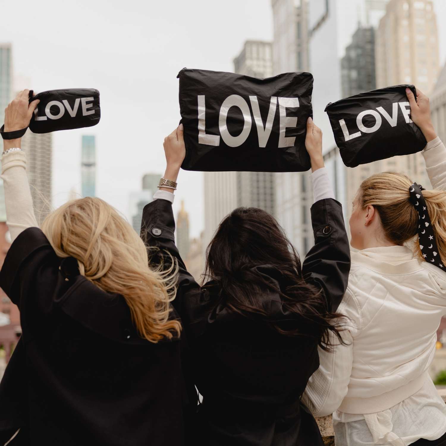 Three women seen from behind holding black zip pouches with the word 'LOVE' in white letters, with a blurred city skyline in the background.