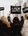 Three women seen from behind holding black zip pouches with the word 'LOVE' in white letters, with a blurred city skyline in the background.