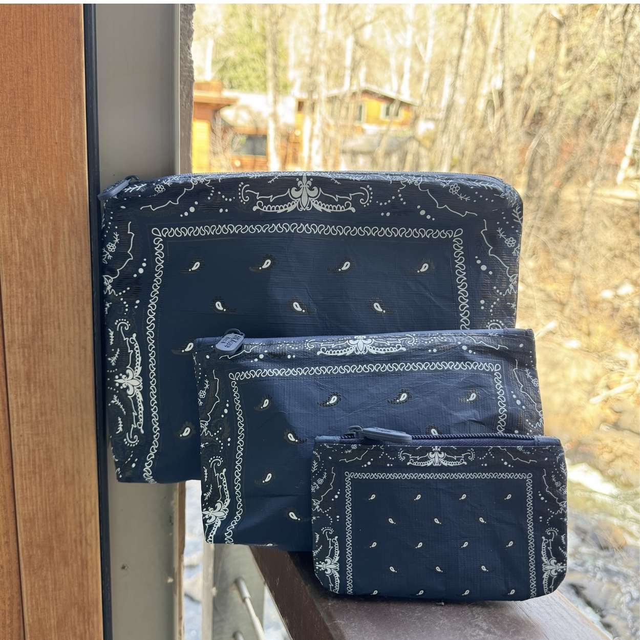Set of three navy blue Bandana patterned pouches on a wooden surface with a window view.
