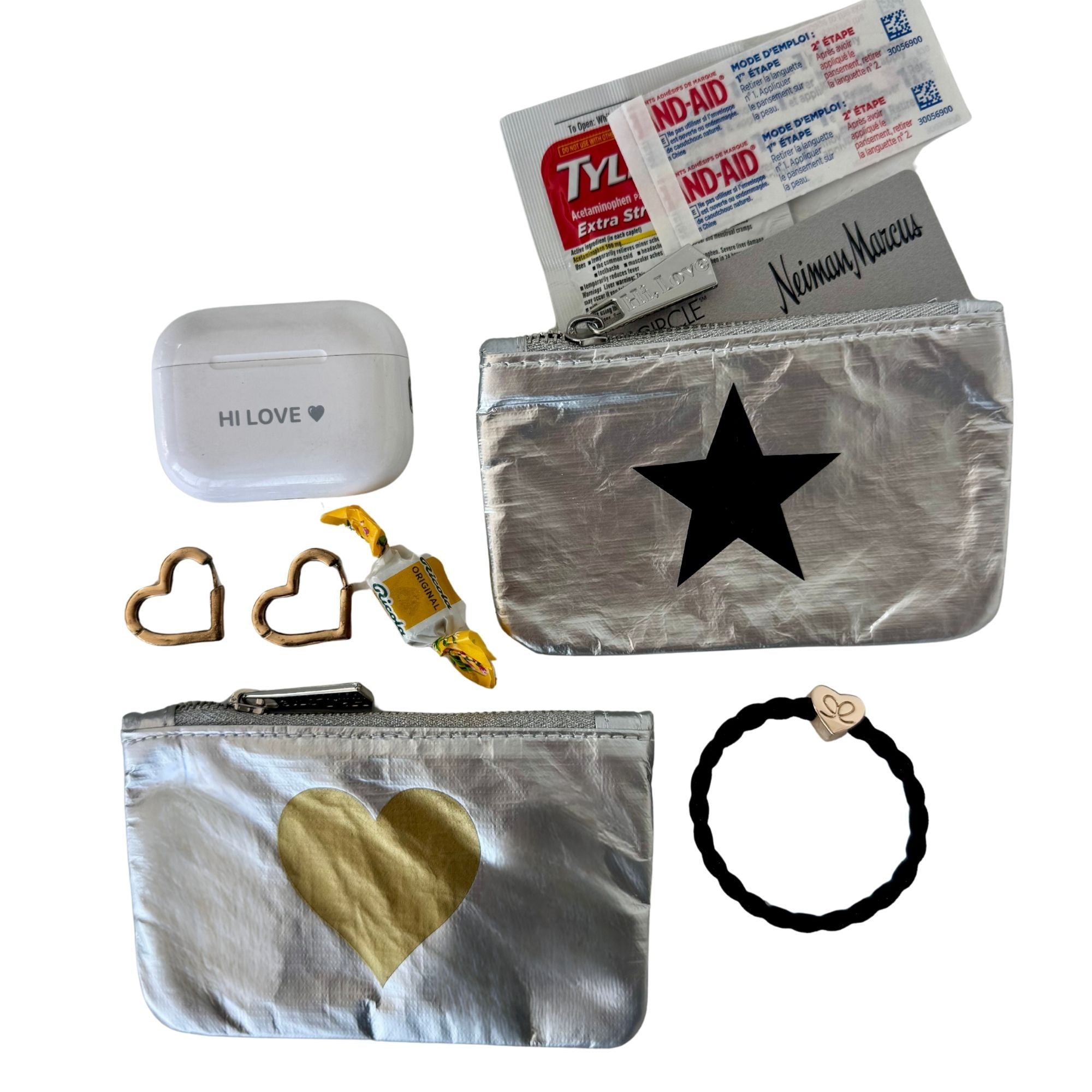 A curated flat-lay of silver metallic zip pouches with heart and star designs, shown alongside accessories including a white AirPods case with 'HI LOVE' printed on it, heart-shaped earings, a mint, credit card, a black braided hair tie with a heart charm, and a package of Tylenol and bandaid.