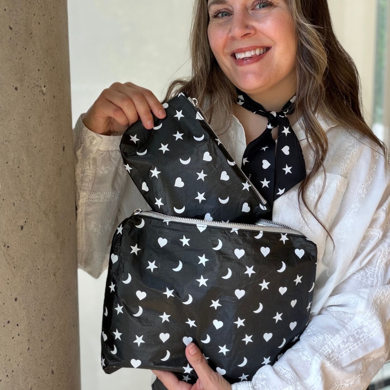 Woman wearing a white blouse holding two zipper pouches in shimmer black with heart moon star print on them.